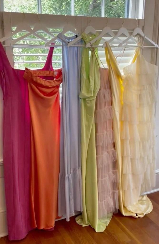 Multicolored satin bridesmaids dresses hanging on a rack in front of windows with sunlight streaming through. Two of the dresses stand out with ruffled designs, adding texture and contrast to the satin collection.