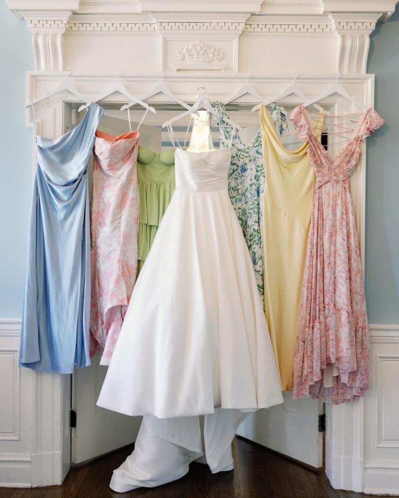 A group of multicolored bridesmaids dresses with floral prints hanging on a wooden rack, accompanied by a white wedding dress in the center. The dresses are light and airy, creating a soft, romantic vibe.