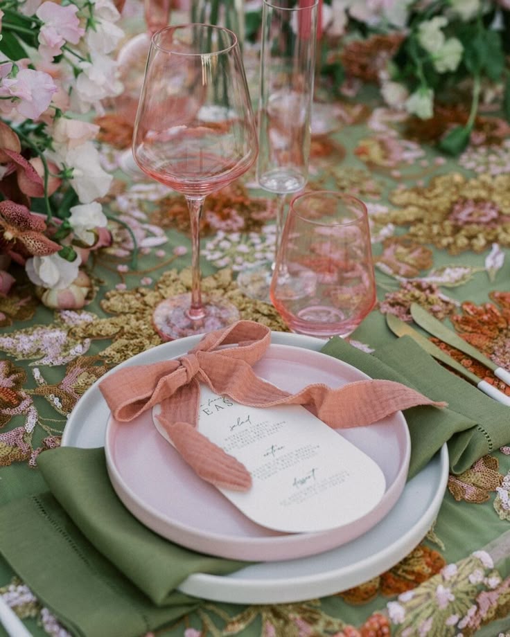 Whimsical garden-inspired wedding place setting with a pink-tinted wine glass, blush and sage green plates, and a pink gauze ribbon tied around a cream-colored menu. The table is decorated with an embroidered green floral linen and surrounded by romantic pastel flowers and gold flatware.
