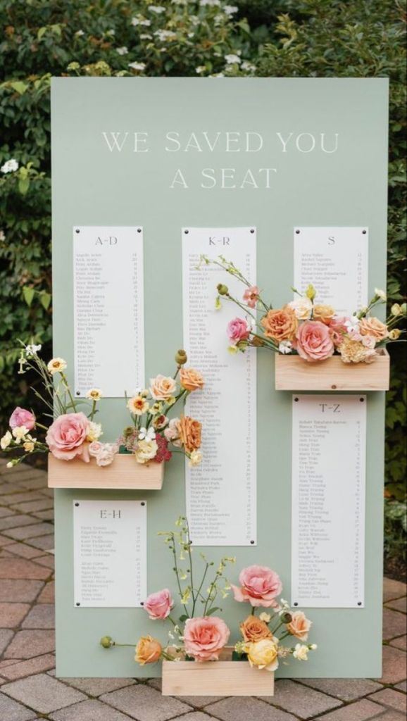 Large sage green wedding seating chart display with the words “We Saved You A Seat” in white serif font. Guest names are organized by last name and printed on six white columns attached to the board. The display is accented with blush, peach, and yellow florals in small wooden planter boxes, set on a paved path with greenery in the background