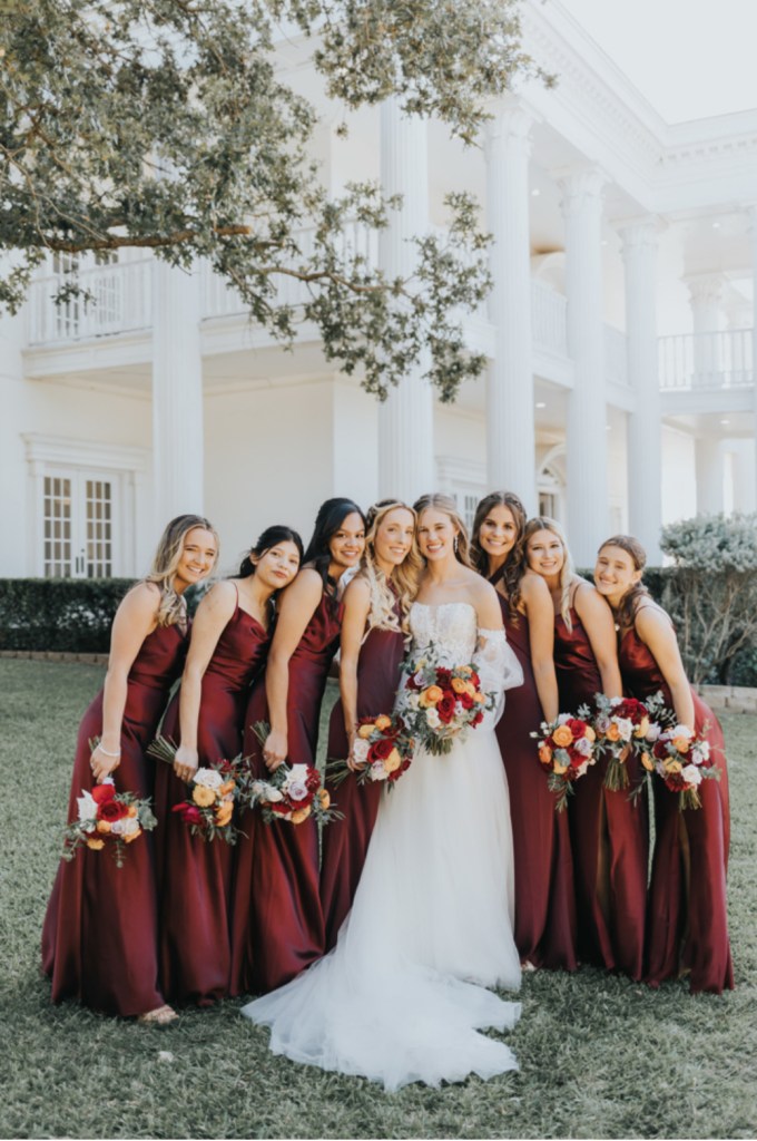 Bride posing with bridesmaids outdoors in matching dark dresses while holding bouquets during an elegant wedding day celebration.