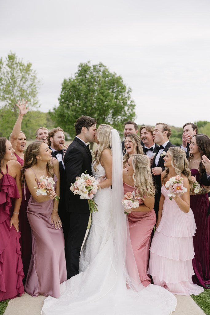 Newlywed couple kissing with their bridal party gathered around them outdoors after the ceremony, capturing an emotional and joyful wedding celebration moment.