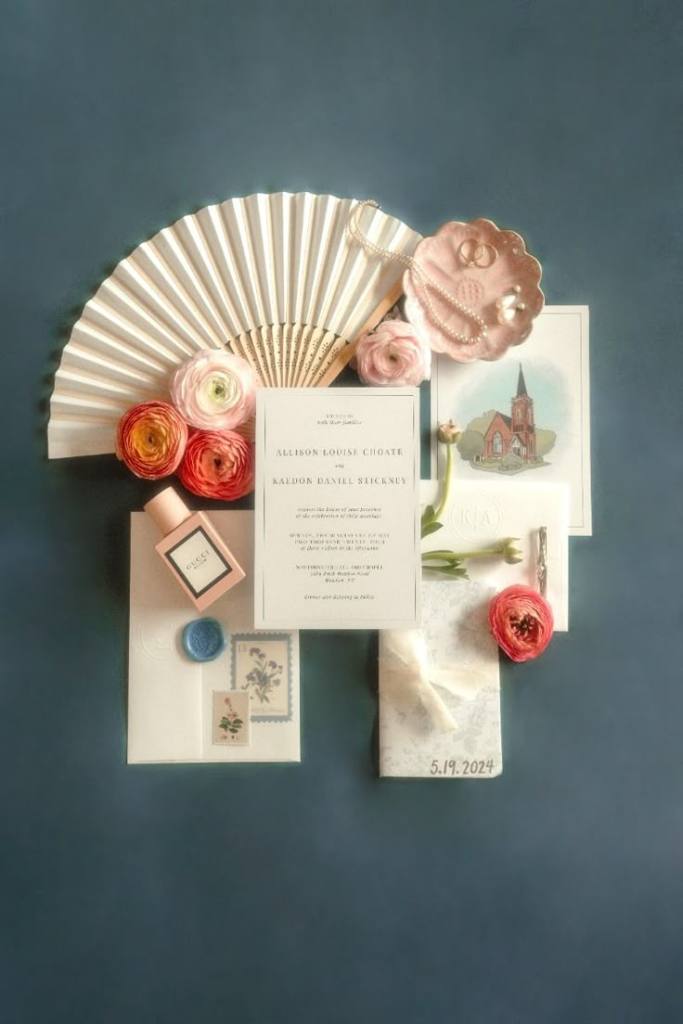 Creative wedding flat lay with invitation suite, fan details, florals, and romantic styling arranged on a dark background.